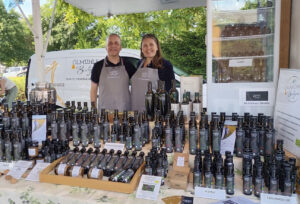 Zwei Personen stehen hinter einem Marktstand, an dem auf Handwerkermärkten und Bauernmärkten verschiedene Ölflaschen mit der Aufschrift "Ölmühle Bröker" und Flaschen mit verschiedenen Ölsorten des Spätsommer Herbst 2022 angeboten werden.