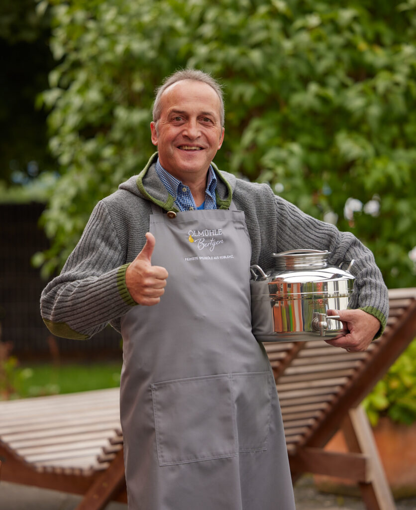 Ein Mann in grauer Schürze und Pullover steht im Freien, hält einen Metalltopf in der Hand und zeigt den Daumen nach oben - bereit für ein spannendes Jahr 2022!.
