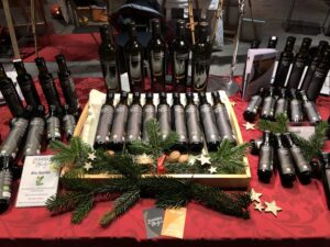 Eine Auslage mit verschiedenen Olivenölflaschen auf einem roten Tischtuch, dekoriert mit Tannenzweigen, Sternen und Nüssen, auf Wochenmärkten und Weihnachtsmärkten im November Dezember 2023.