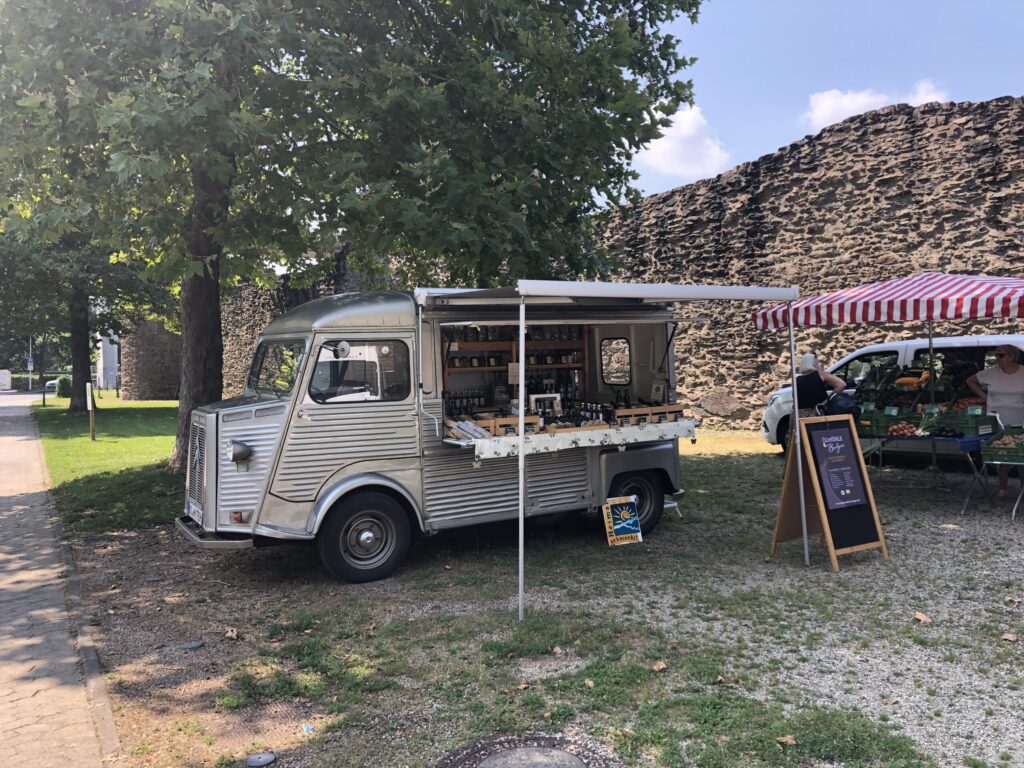 Ein silberner Foodtruck mit offenem Servicefenster parkt an einer Steinmauer neben einem Marktstand unter einem rot-weiß gestreiften Vordach auf dem belebten Sondermarkt 2024.