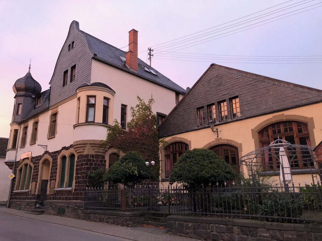 Ein großes, historisches Gebäude in der Eifel mit Stein- und Stuckwänden, Bogenfenstern, einem Türmchen und einem Gartenbereich davor, an einer ruhigen Straße in der Abenddämmerung - perfekt für diejenigen, die sich in eine ruhige Umgebung umziehen möchten.