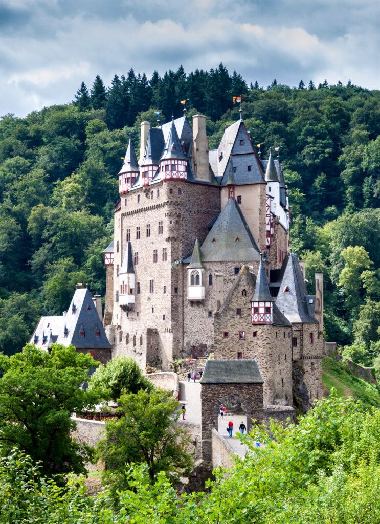 Eine mittelalterliche Steinburg mit spitzen Türmen thront auf einem Hügel, umgeben von grünen Bäumen und Wäldern, und regt unter einem teilweise bewölkten Himmel zu philosophischen Gedanken an.