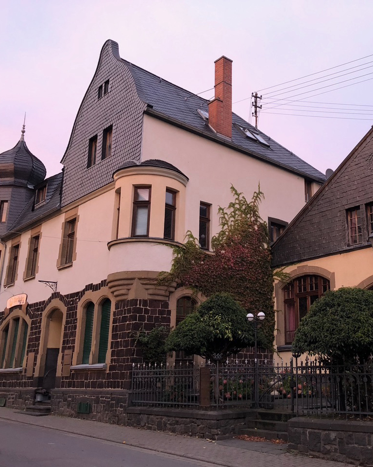 Ein dreistöckiges historisches Gebäude mit steilem Dach, bogenförmigen Fenstern und Efeu an der Steinfassade, fotografiert bei Sonnenuntergang an einer ruhigen Straße.