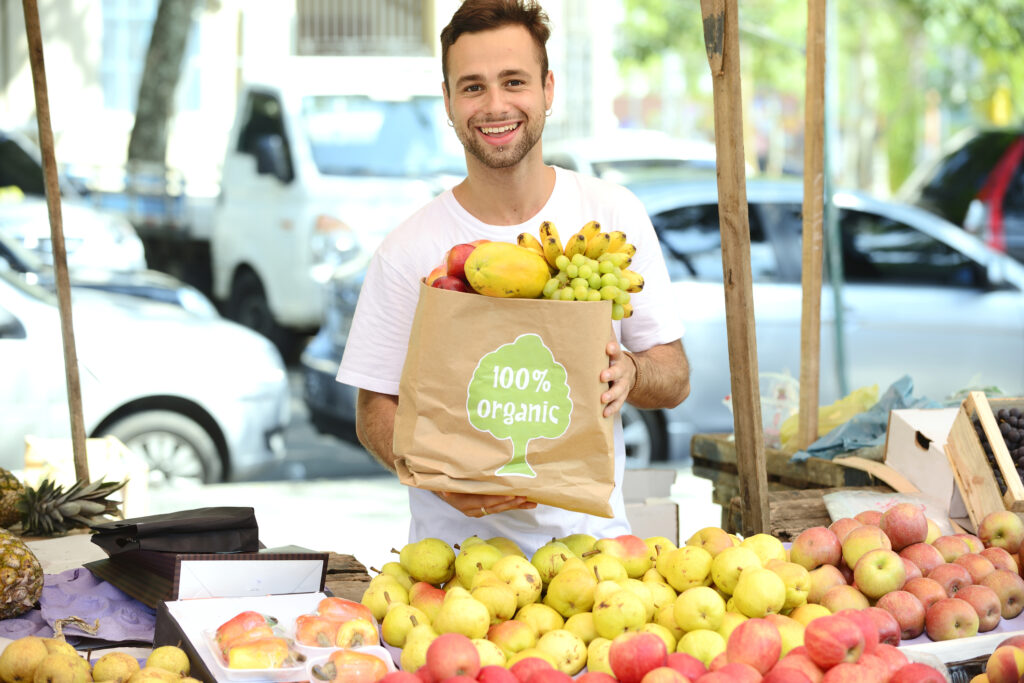 Eine Person auf dem Andernacher Markt hält eine mit Obst gefüllte Papiertüte mit der Aufschrift "100% Bio" in der Hand. Sie steht hinter einem Tisch mit Äpfeln, Birnen und anderen frischen Produkten auf dem belebten Wochenmarkt.